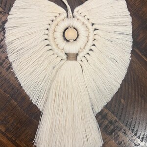 May include: A white macrame angel ornament with a wooden bead in the center. The angel has a long tassel hanging from the bottom.