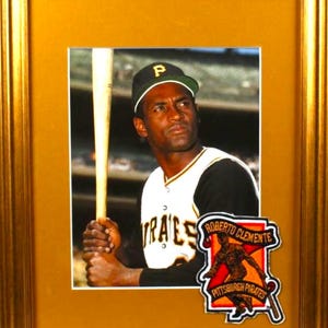 May include: Framed photograph of Roberto Clemente in a Pittsburgh Pirates uniform, holding a baseball bat. The image includes a team logo patch and is set in a gold-colored frame.