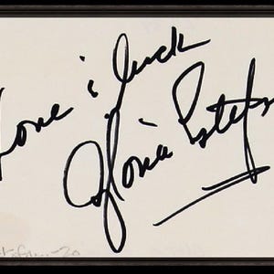May include: Framed artwork featuring the handwritten words "Love i luck" and the signature "Gloria Estefan" in black ink on a cream-colored background. The artwork is set within a dark brown frame.