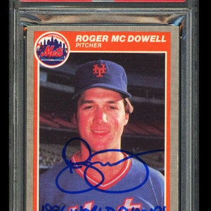 May include: A baseball card featuring a photo of Roger McDowell, a pitcher for the New York Mets. The card is signed by McDowell and includes the text "1986 World Champs" and "Fleer".