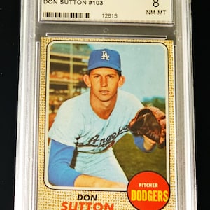May include: A 1968 Topps Don Sutton #103 baseball card graded NM-MT 8 by GRADEMYCARDS.COM. The card features a player in a light blue Dodgers uniform and blue cap, with the text DON SUTTON and PITCHER DODGERS printed on a textured tan background.