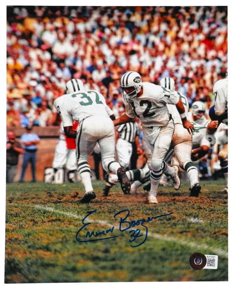 May include: A black and white photo of a football game with three players in green and white uniforms. The player in the center is wearing number 12 and has signed the photo "Emmitt  Boozer 32".