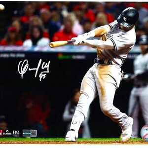 May include: A baseball player in a white and grey uniform swings a bat. The player is wearing a black baseball cap with the letters "NY" on it. The player is in a stadium with a crowd of people in the background. The player's signature is in the lower left corner of the image. The signature reads "C-Jay 95".
