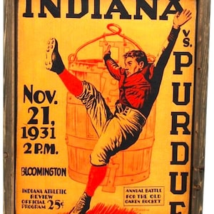 May include: Framed vintage poster for the Indiana vs. Purdue football game, dated November 21, 1931. The artwork features a player reaching for a wooden bucket, with text in black and red on a yellow background. The poster is in a wooden frame.