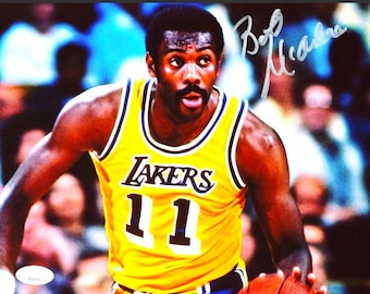Pode incluir: Um close-up de um jogador de basquete vestindo uma camisa amarela e azul dos Lakers com o número 11 na frente. O jogador está driblando uma bola de basquete e tem uma expressão séria no rosto. A assinatura do jogador está no canto superior direito da imagem.