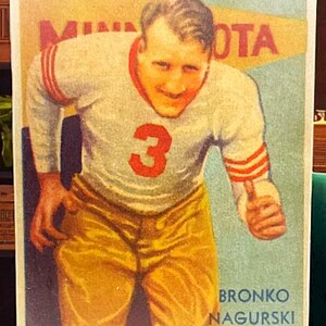 May include: Vintage print featuring a football player in a white jersey with red stripes and the number 3. The player is wearing gold pants and is set against a backdrop with the word "MINNESOTA". The print also includes the name "BRONKO NAGURSKI".