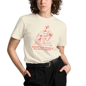 Könnte beinhalten: Ein weißes T-Shirt mit einer roten Illustration einer Frau, die auf einem Löwen sitzt. Der Text "burdened by the anchors of self-respect & dignity" ist in rot unter der Illustration aufgedruckt.