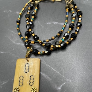 May include: A layered bracelet with black, faceted beads, gold-coloured spacers, and a cream-coloured rectangular pendant. The pendant has black markings and gold-coloured floral accents. The bracelet has a gold-coloured chain clasp.