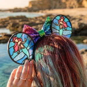 May include: A colorful headband featuring two round ears with stained-glass style mermaid designs. The headband has a sequined bow with rainbow colors. The image is taken outdoors near the ocean.