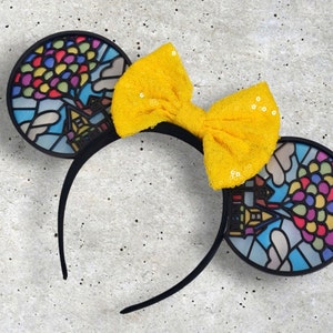 May include: Black mouse ears with a stained glass design of a house and balloons. A yellow bow with sequins is attached to the headband.