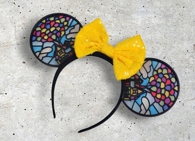 May include: Black mouse ears with a stained glass design of a house and balloons. A yellow bow with sequins is attached to the headband.