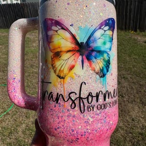 May include: A glittery pink and white travel mug with a handle. The mug features a colorful butterfly design with the words "Transformed by God's Love". The mug is designed to keep beverages hot or cold.