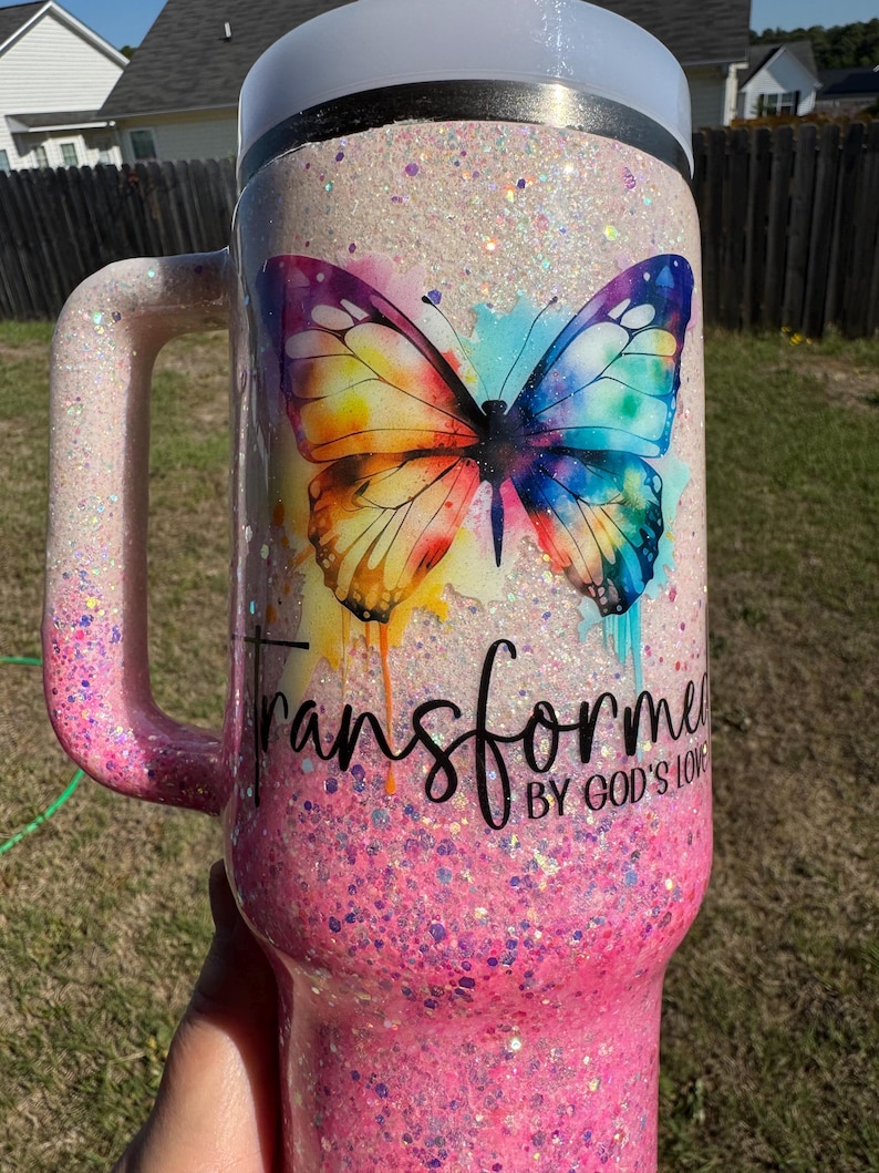 May include: A glittery pink and white travel mug with a handle. The mug features a colorful butterfly design with the words "Transformed by God's Love". The mug is designed to keep beverages hot or cold.