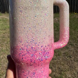 May include: A large, glitter-covered tumbler with a handle. The cup transitions from white to pink, with a gradient of glitter. The handle is pink. The lid is white. The tumbler is held by a hand.