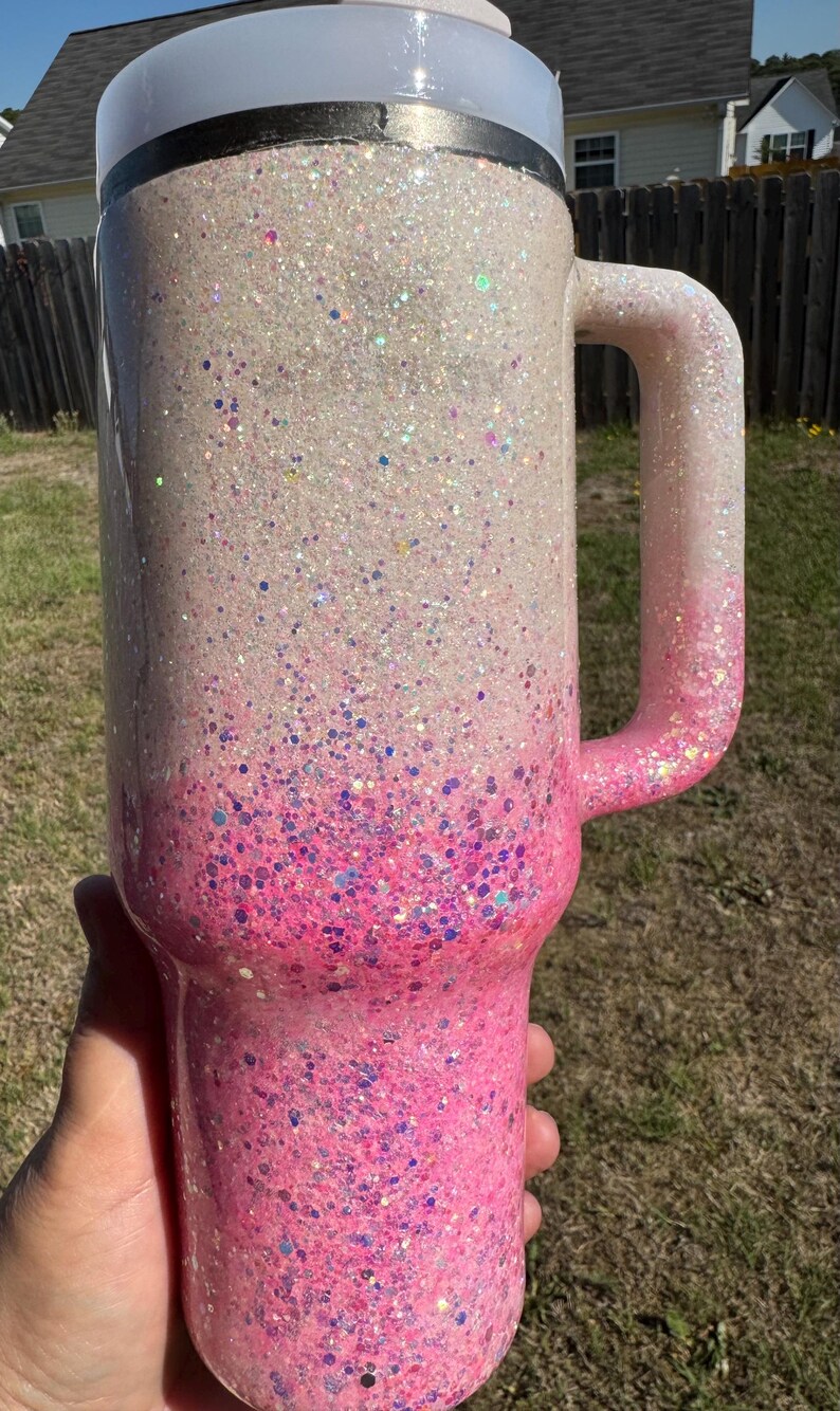 May include: A large, glitter-covered tumbler with a handle. The cup transitions from white to pink, with a gradient of glitter. The handle is pink. The lid is white. The tumbler is held by a hand.