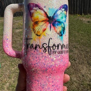 May include: A glittery pink and white tumbler with a handle. The tumbler features a colorful butterfly design and the words "Transform" and "By God's Love". The handle and base are pink, and the top is white.
