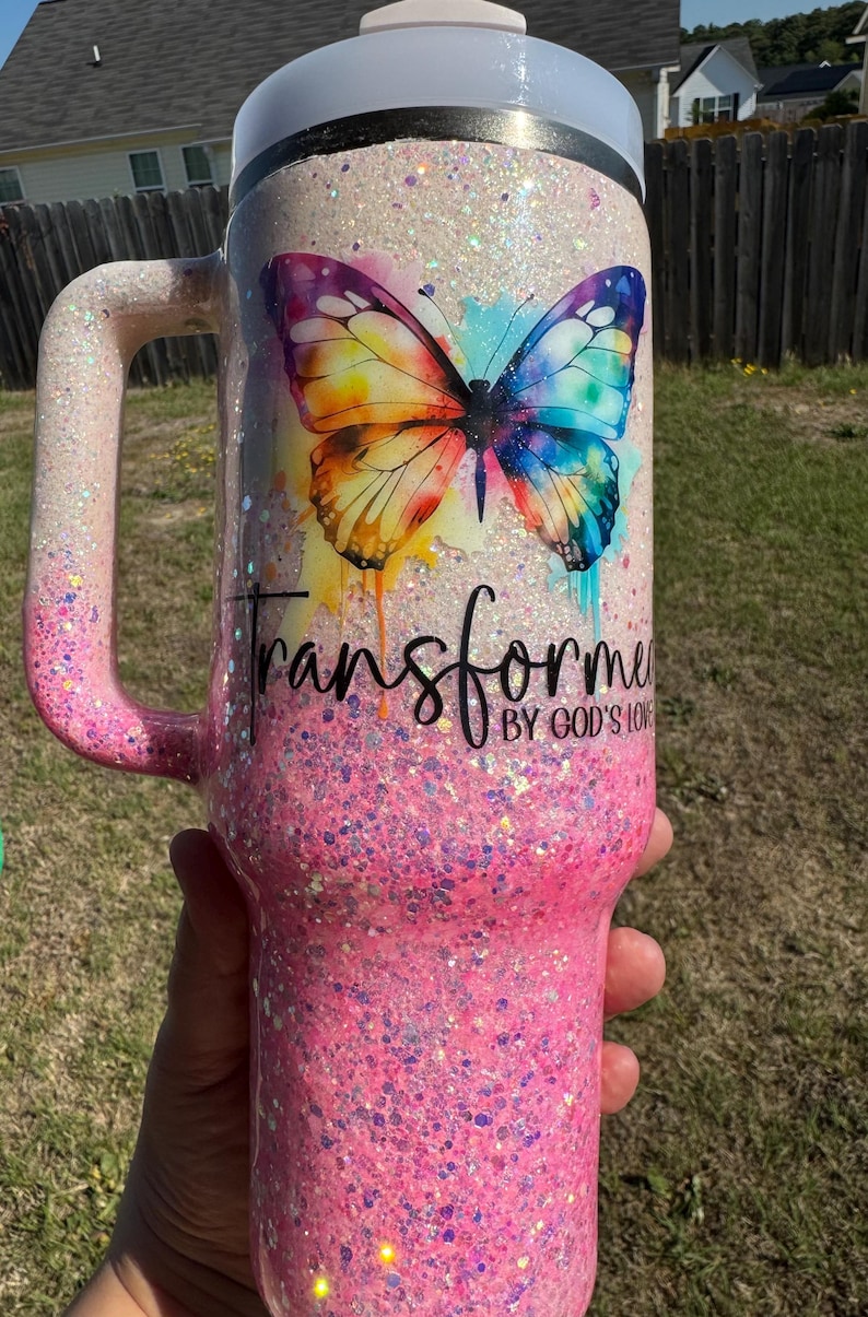 May include: A glittery pink and white tumbler with a handle. The tumbler features a colorful butterfly design and the words "Transform" and "By God's Love". The handle and base are pink, and the top is white.