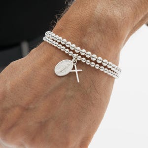 May include: A silver beaded bracelet with two strands, featuring a pendant with a religious image and a small cross charm. The bracelet is worn on a wrist.