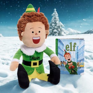 May include: A plush Edwin doll in a green elf costume, black belt and shoes. The doll has brown hair and a smiling face. Next to it is a book titled "elf" with a cartoon illustration.