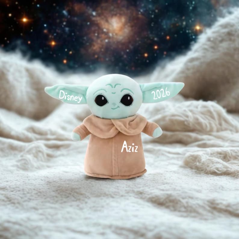 May include: A soft toy of a character with large black eyes, mint green skin, and large ears. The toy is wearing a tan robe. The word "Disney" is on one ear, "2026" on the other, and "Aziz" on the robe.