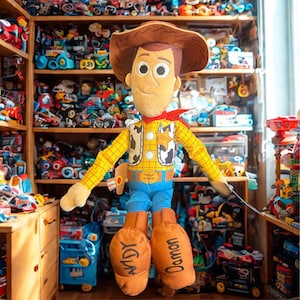 Puede incluir: Mu&ntilde;eco de peluche de Woody de la franquicia Toy Story. El mu&ntilde;eco presenta un sombrero de vaquero marr&oacute;n, una camisa amarilla con un chaleco con estampado de vaca, pantalones azules y botas naranjas con los nombres "Andy" y "Damon" impresos. Un pa&ntilde;uelo rojo est&aacute; atado alrededor del cuello.