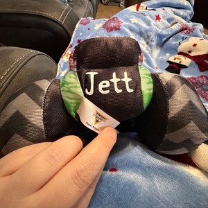May include: A plush toy with the name "Jett" in white lettering on a black background. The toy has black and gray patterned sides and green and red accents. A "Monster Jam" tag is visible.