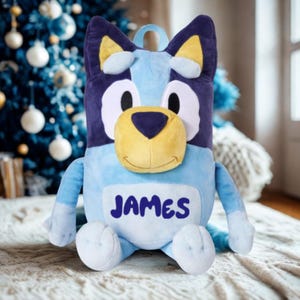 May include: A light blue dog-shaped cushion with the name "JAMES" embroidered on its front. The soft toy has dark blue ears, a yellow nose, and a smiling face. It is placed on a textured surface, with a Christmas tree visible in the background.