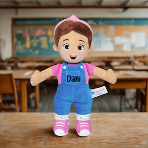 Puede incluir: Muñeca de peluche de una niña con una camisa rosa, un peto azul con el nombre "Dani" y zapatos rosas. La muñeca tiene el pelo castaño y una diadema rosa. Una etiqueta dice "Ms. Rachel".