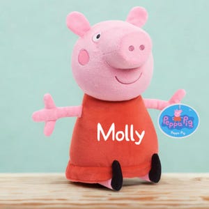 May include: A plush toy of the character Molly from Peppa Pig. The pink pig has a red dress with the name "Molly" printed on it. The toy has a round tag with the Peppa Pig logo.