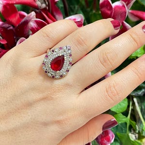 May include: A teardrop-shaped ring with a large red gemstone surrounded by smaller clear and red gemstones. The ring is set in gold.