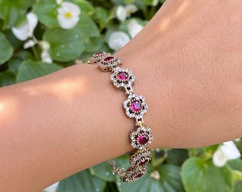 Pode incluir: Uma pulseira de prata com um design de corrente com detalhes de pedras preciosas vermelhas em um padrão floral.