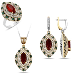 Puede incluir: Un conjunto de joyas de plata, oro y rojo con un anillo, un collar y pendientes. El conjunto presenta una piedra preciosa roja rodeada de piedras preciosas blancas y verdes más pequeñas.