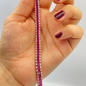 May include: A rose gold bracelet with alternating rows of rectangular, deep pink gemstones and small, clear gemstones. The bracelet is held against a light background, showcasing its intricate design and vibrant colors.