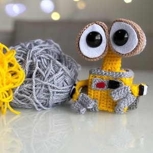 Puede incluir: Un juguete robot de crochet amarillo con grandes ojos negros y un botón rojo en el pecho. El robot está sentado junto a una bola de hilo gris.