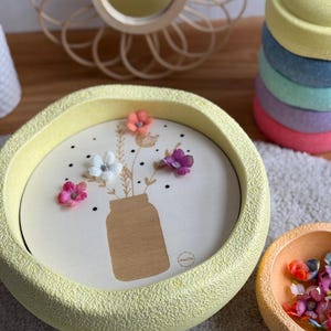 Könnte beinhalten: Ein gelber, strukturierter, runder Dekoteller mit Holzeinsatz, der eine Gläser-Illustration und bunte Blumen zeigt. Eine kleinere orangefarbene Schale mit Blumen befindet sich neben dem Tablett. Ein Stapel bunter runder Objekte befindet sich im Hintergrund.