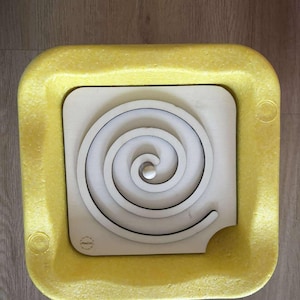 Könnte beinhalten: Ein Holz-Spiral-Labyrinth-Puzzle mit einer silbernen Kugel, eingebettet in einen quadratischen, hellfarbigen Holzrahmen. Der Rahmen ist von einem gelben, strukturierten Rand umgeben. Das Puzzle ist für die Hand-Augen-Koordination und Problemlösung konzipiert.