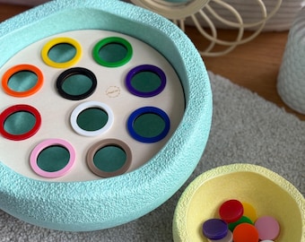 May include: A light blue wooden bowl with a textured surface. The bowl has a circular opening with 12 colorful plastic rings. A smaller yellow bowl with a textured surface contains the same rings.