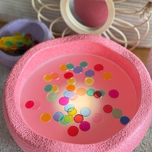 Könnte beinhalten: Ein rosa, sensorischer Behälter aus Plastik, gefüllt mit bunten, durchsichtigen Kreisen. Der Behälter hat eine strukturierte Oberfläche und ist mit Wasser gefüllt.