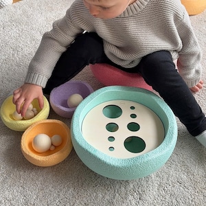 Könnte beinhalten: Ein Kind spielt mit einem pastellfarbenen Holzspielzeug mit verschiedenen Lochgrößen und passenden Bällen. Das Spielzeug befindet sich auf einem grauen Teppich.