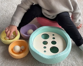 May include: A child plays with a pastel-colored wooden toy with a variety of sized holes and matching balls. The toy is on a gray carpet.