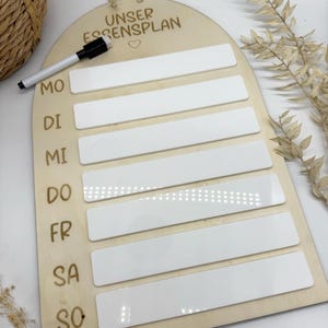Könnte beinhalten: Wöchentlicher Essensplaner aus Holz mit dem deutschen Text "UNSER ESSENSPLAN" und einem Herzsymbol. Der Planer hat sieben weiße rechteckige Abschnitte für jeden Wochentag, beschriftet mit MO bis SO. Ein schwarz-weißer Marker ist enthalten.
