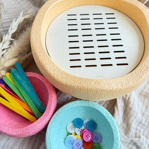 Könnte beinhalten: Ein Set aus drei strukturierten Schalen in Pastellfarben. Die größte Schale ist beige mit einem weißen, geschlitzten Einsatz. Eine rosa Schale enthält bunte Bastelstäbchen und eine blaue Schale enthält verschiedene Knöpfe. Die Artikel sind auf einem neutralen Stoff angeordnet.