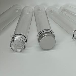 Könnte beinhalten: Fünf transparente Plastikröhrchen mit silbernen Deckeln. Die Röhrchen sind zylindrisch und haben einen schwarzen Streifen um die Oberseite.