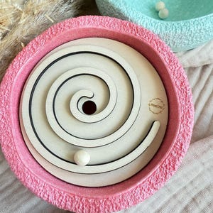 Könnte beinhalten: Eine rosa, strukturierte Schale mit einem Holz-Spiral-Labyrinth und einer weißen Kugel. Das Labyrinth hat schwarze Linien und ein zentrales Loch. Im Hintergrund befindet sich eine hellblaue Schale mit weißen Kugeln. Thema: Kinderspielzeug.