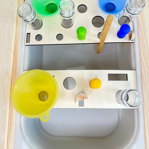 Könnte beinhalten: Ein Holztablett mit einem weißen Waschbecken darunter. Das Tablett hat Löcher für Reagenzgläser und Trichter. Es gibt drei Trichter, einen grünen, einen blauen und einen gelben. Außerdem gibt es zwei Holzlöffel, einen Holzstab und ein paar bunte Silikonobjekte.