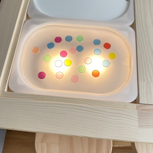Könnte beinhalten: Ein sensorischer Spieltisch mit einer weißen Kunststoffschale, die von unten beleuchtet wird und mit farbigen, durchscheinenden Scheiben gefüllt ist. Der Tisch besteht aus hellem Holz, mit einem passenden Deckel und einem kleinen Holzschemel.