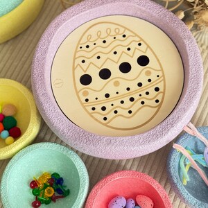 Könnte beinhalten: Ein Set pastellfarbener Schalen mit unterschiedlichen Texturen und Designs. Die Schalen sind mit verschiedenen kleinen Gegenständen gefüllt, wie z. B. bunten Pompons, Stecknadeln und gesprenkelten Eiern. Die größte Schale hat ein hölzernes Eidesign mit schwarzen Punkten und Wellenlinien.
