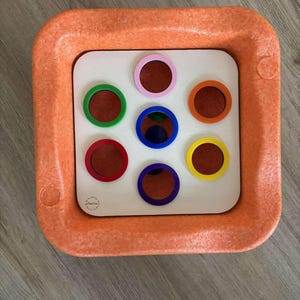 Könnte beinhalten: Ein orangefarbenes quadratisches sensorisches Spielzeug mit einem weißen Mittelteil mit sieben farbigen Kreisen. Die Kreise sind rot, grün, rosa, orange, blau, lila und gelb. Das Spielzeug hat einen strukturierten orangefarbenen Rahmen.