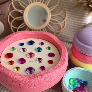 Könnte beinhalten: Eine rosa Schale mit weißem Boden, gefüllt mit bunten Edelsteinen. Die Schale steht auf einer gewebten Oberfläche. Es gibt eine kleinere blaue Schale mit Edelsteinen darin.