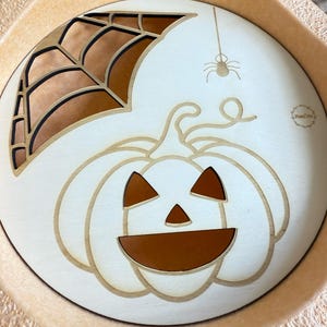 Könnte beinhalten: Runder Halloween-Holztablett mit geschnitztem Kürbisgesicht, Spinne und Spinnennetz-Design. Der Kürbis hat ein braunes Gesicht und ist von hellem Holz umgeben. Das Tablett hat einen hellbraunen Rand.